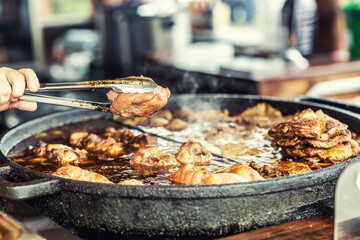 Hand holding a piece of meat taken from a large frying pan in kitchen pliers