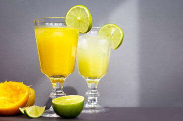 two glasses with a refreshing citrus drink or lemonade or alcoholic cocktail on grey background, a pair of vintage goblets among sliced lemons, oranges and limes, copy space