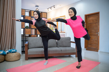 two women wearing a headscarf smiling while doing lunges movements while exercising indoors together at home