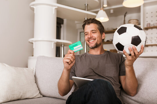 Young Soccer Fan Man Watching Football Game On Television At Living Room Couch. He Holds A Credit Card And A Digital Tablet For Online Betting.