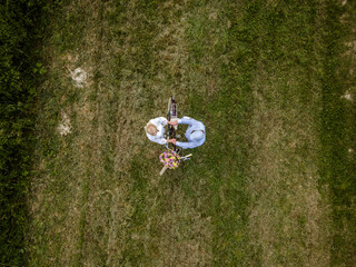 aerial drone shot, pic of blonde woman with white dress and her boyfriend or husband posing with bicycle with beautifully decorated flower basket in nature, nature concept, flowers, delivery service