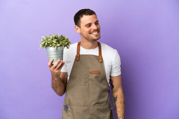 Brazilian Gardener man holding a plant over isolated purple background looking to the side and smiling