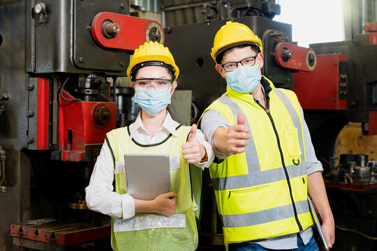 Asian Woman And Man Wearing A Mask Holding A Tablet And Show Thumb Up In An Industrial Factory. Professional Technicians Are Holding A Tablet To Control Work In Industrial Plants.