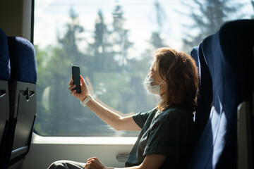 Young happy woman in protective mask sitting in high speed train chatting online using smartphone