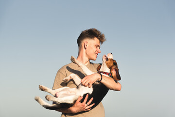 Happy young man holds his funny beagle dog, shot against clear blue sky with copy space. Lifestyle with pets, happy puppy owner, pedigree dog - Powered by Adobe