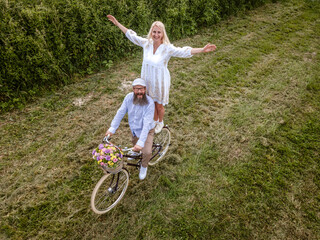 aerial drone shot, pic of blonde woman with white dress and her boyfriend or husband posing with bicycle with beautifully decorated flower basket in nature, nature concept, flowers, delivery service