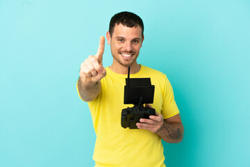 Brazilian man holding a drone remote control over isolated blue background showing and lifting a finger