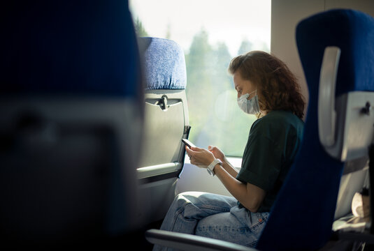 Young Woman In A Protective Mask Sitting In A Train With A Smartphone In Her Hands. Taking Care Of Health And Travel During A Pandemic