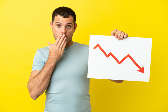 Brazilian man over isolated purple background holding a sign with a decreasing statistics arrow symbol with surprised expression