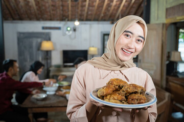 smiling veiled woman carrying a plate of fried chicken with family members eating together in the dining room as a background