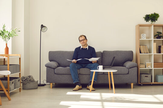 Relaxed Mature Man Sitting On Sofa In The Living-room, Holding Papers And Looking At Camera. Senior Person Reading Pension Plan Or Going Over Business Documents While Doing Paperwork At Home