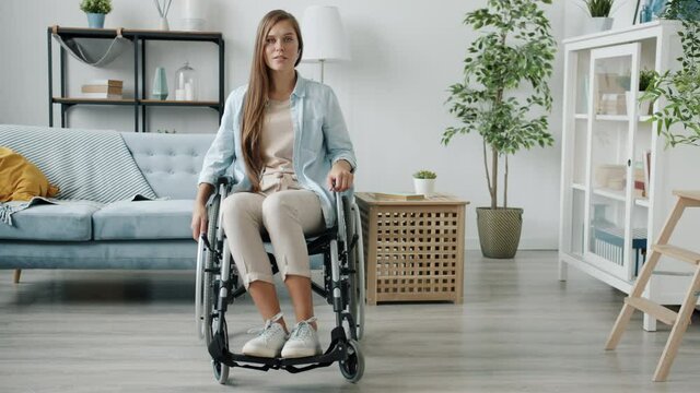 Slow motion portrait of beautiful paraplegic lady sitting in wheelchair in modern room and looking at camera. Physical impairment and house concept.