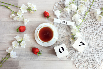 Calendar for July 7 : cubes with the numbers 0 and 7, the name of the month of July in English, a cup of tea on a gray openwork napkin, scattered flowers of bluebells and strawberries, top view