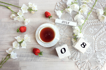 Calendar for July 8 : cubes with the numbers 0 and 8, the name of the month of July in English, a cup of tea on a gray openwork napkin, scattered flowers of bluebells and strawberries, top view