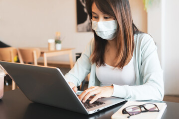 Asian woman working on computer and wearing medical mask on table in coffee shop during the coronavirus crisis. Business, health care and new normal lifestyle concept.