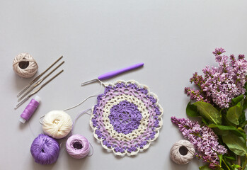 Fototapeta na wymiar Spring needlework and hobby. Hand crocheting a delicate round lace doily. Sprig of blooming lilacs on a table. Handicrafts and DIY concept. Empty space for text on top. Flat lay, copy space, close up