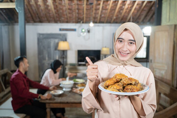 woman in veil carrying a plate of fried chicken with thumbs up in the background family members eating together in the dining room