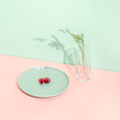 Minimal summer fruit concept with red organic cherry on light green plate and glass vase with grass  against orange background. Vegan plant based nutrition.