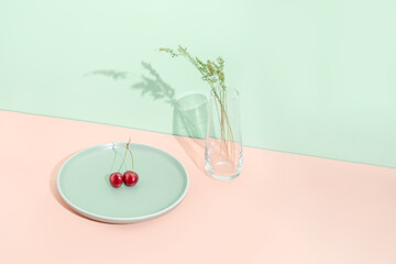 Minimal summer fruit concept with red organic cherry on light green plate and glass vase with grass  against orange background. Natural healthy diet and fresh food idea.