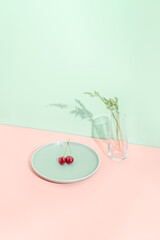 Minimal sunny summer fruit concept with red organic cherry on light green plate and glass vase with grass on orange background. Natural healthy diet and fresh food idea. Vegan plant based nutrition.