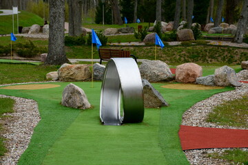 A close up on a metal loop being a part of a mini golf course seen next to some boulders, rocks, or stones, gravel finish, grass surface and blue flags showing where individual holes are 
