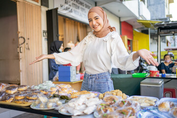 smiling beautiful girl in hijab with hand gestures offering a variety of fried foods by the side of the road