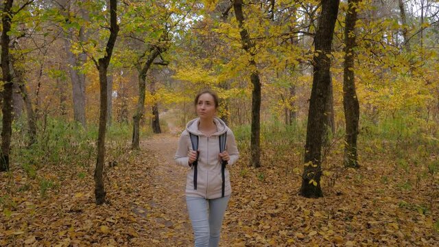 Front view of young positive woman with backpack walking in autumn park, forest and looking around - wide angle steadicam shot. Active outdoor lifestyle, leisure time, freedom and adventure concept