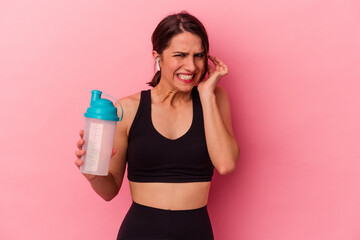 Young caucasian woman drinking a protein shake isolated on pink background covering ears with hands.