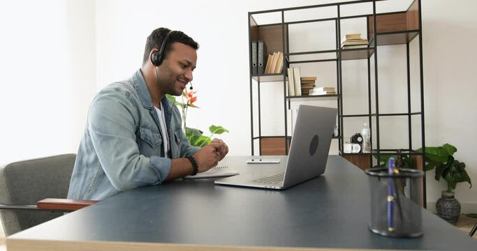 Smiling young indian man in headset with headphones and laptop at the table makes notes in a notebook. Talking, meeting online or e-education. Customer support