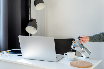 An unrecognizable female hand pours coffee into a cup standing on a desktop.