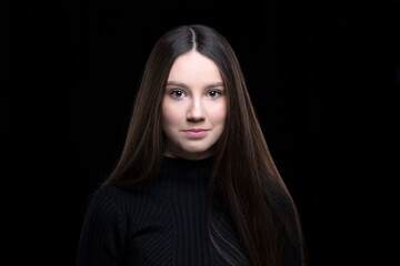 Fototapeta premium Beautiful girl with dark long hair on a black background. Seventeen-year-old woman. Proper facial features.