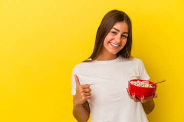 Young caucasian woman holding cereals isolated on yellow background smiling and raising thumb up