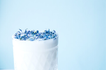 Beautiful crystal glass with milk and blue flower petals on blue background. Abstract background. Floral pattern of blue flower petals on white background