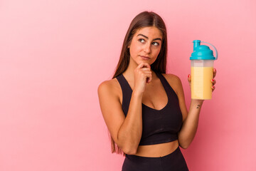 Young caucasian woman holding a protein shake isolated on yellow background looking sideways with...
