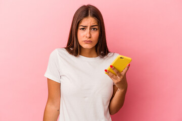 Young caucasian woman holding a mobile phone isolated on pink background shrugs shoulders and open...