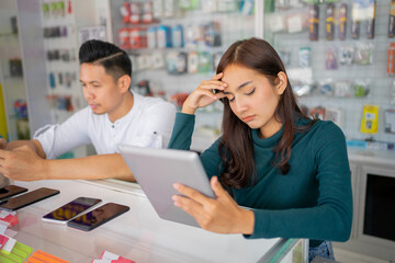 a dizzy woman holding her head while using a tablet beside a man sitting using a cell phone in a smartphone shop