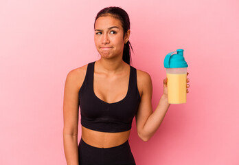 Young Venezuelan woman drinking a protein shake isolated on pink background confused, feels...