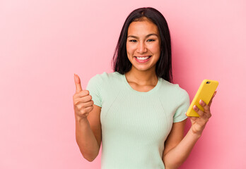 Young Venezuelan woman holding a mobile phone isolated on pink background smiling and raising thumb...