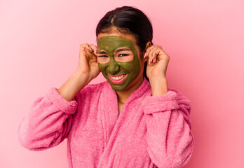 Young Venezuelan woman wearing a bathrobe and facial mask isolated on pink background covering ears with hands.