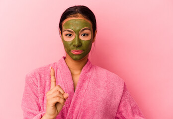 Young Venezuelan woman wearing a bathrobe and facial mask isolated on pink background showing number one with finger.