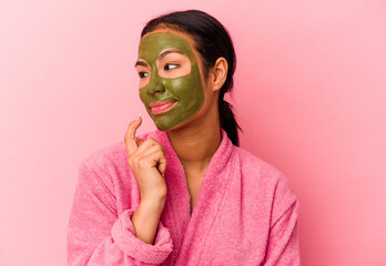 Young Venezuelan woman wearing a bathrobe and facial mask isolated on pink background relaxed thinking about something looking at a copy space.