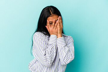 Young Venezuelan woman isolated on blue background blink at the camera through fingers, embarrassed covering face.