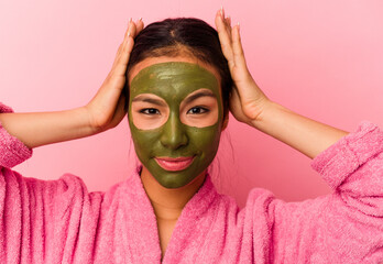 Young venezuelan woman wearing a bathrobe and facial mask isolated on pink background