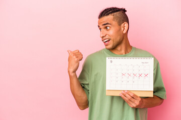 Young venezuelan man holding a calendar isolated on pink background points with thumb finger away, laughing and carefree.