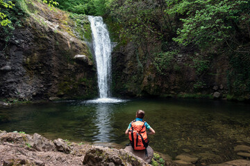 Obraz premium Child boy schoolboy with hiking backpack sitting alone in forest near lake and watching waterfall. Children's exploration and curiosity of nature, adventure learning and outdoor education