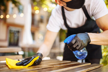 A waiter in a black protective mask wipes tables with a disinfectant before opening. Dust cleaning agent. Wipes the table with a rag in a restaurant.