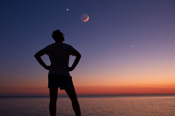 Silhouette of a man looking at the Moon and stars over sea ocean horizon.