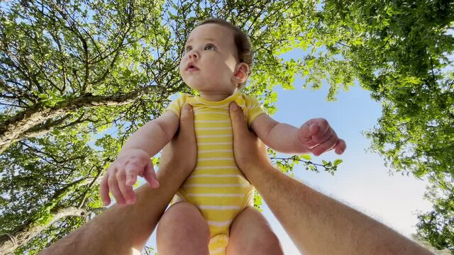 POV shot dad holds small happy smiling cute child at arm length above him, playing together in nature with trees and blue sunny sky in background, filmed first person, carefree family enjoys weekend.