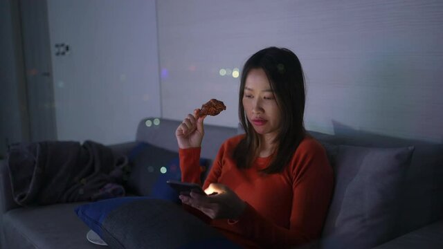Asian woman sitting on sofa in living room at apartment and eating fried chicken for dinner with using mobile phone at night. Female enjoy city life indoor lifestyle with wireless technology at home.