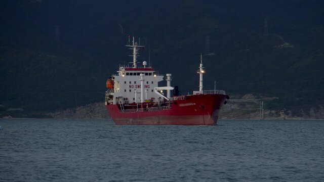 The red cargo ship is at anchor. Night time. Against the background of the shore. Water waves. Sea harbor.